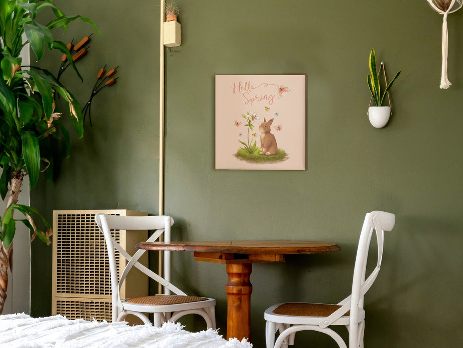12x12 canvas with Hello Spring Easter print with a bunny and butterflies on green bedroom wall hanging over a wooden table with two white chairs surrounded by house plants and a white bed mattress