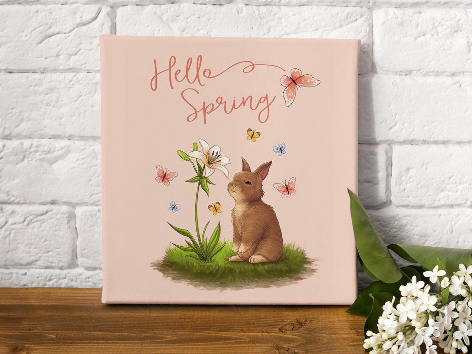 12x12 canvas with Hello Spring Easter print with a bunny and butterflies on wooden table next to white flowers in front of a white brick wall