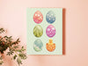 8x10 canvas with colorful easter artwork of rows of eggs and a baby chick on an orange wall next to a house plant