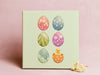 12x12 canvas with colorful easter artwork of rows of eggs and a baby chick in front of a pink wall with little white flowers in front