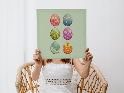 woman in white dress sitting in a wicker chair holding a 12x12 canvas with colorful easter artwork of rows of eggs and a baby chick in a living room with white walls and curtains