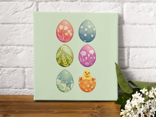 12x12 canvas with colorful easter artwork of rows of eggs and a baby chick on a wooden table next to a small bunch of white flowers in front of a white brick wall