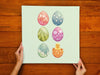 two hands holding a  12x12 canvas with colorful easter artwork of rows of eggs and a baby chick on a wooden table