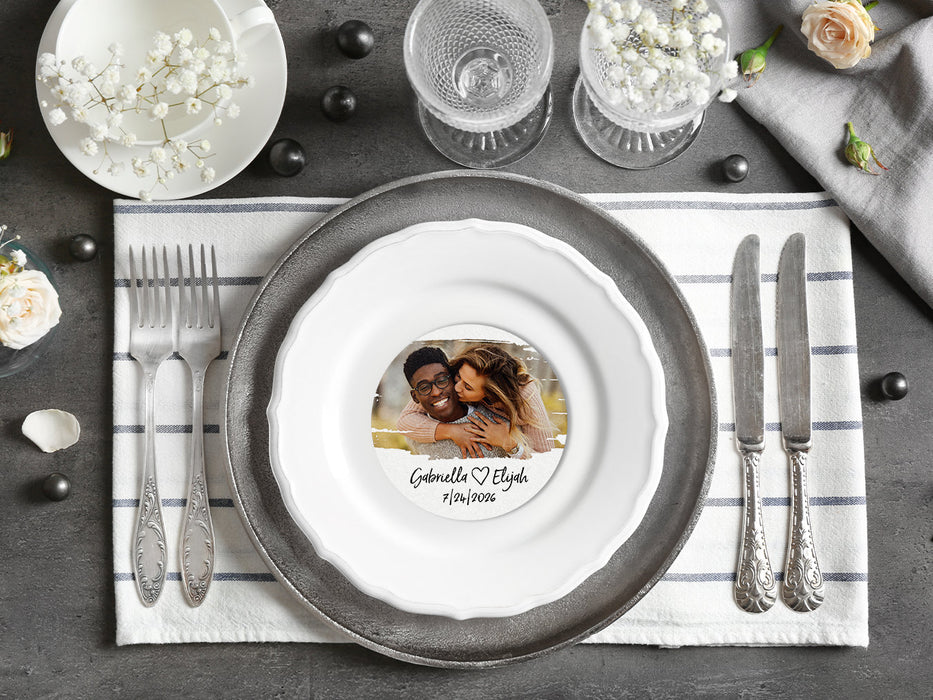 A coaster is shown as a part of a place setting with flowers and silverware. Coaster shown is customizable. Coasters are designed with custom photo, text, and brushed elements. Coaster text reads Gabriella &lt;3 Elijah, 7/24/2026