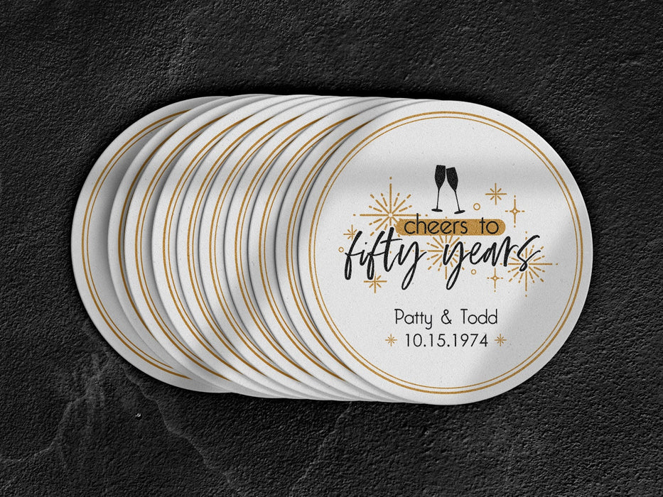 A stack of coasters are shown spread out on a black surface. Coasters shown are customizable. Coasters are designed with gold sparkly elements, wine glasses, and the words Cheers to twenty-five years, custom names, and custom date.