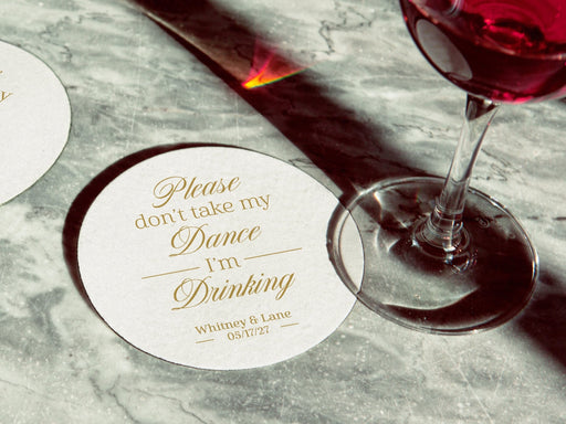 Coaster is shown with a wine glass on top of it and another off to the side. Coasters say Please don't take my dance, I'm drinking with wedding couple's names and wedding date on the bottom.