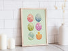 wooden frame with pastel easter art print of decorated eggs with a baby chick on a white kitchen counter top surrounded by candles and a potted plant