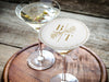 One coaster is shown being used as drink cover on top of a martini glass. Coasters feature a custom floral monogram design with two initials and floral decoration in the middle and a border with a wedding date on the side.