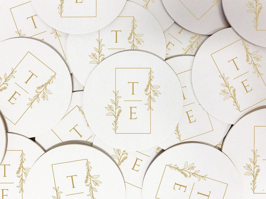 A pile of custom round coasters is shown. Coasters feature a personalized floral monogram design with first name initials surrounded by a floral frame.