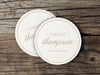 A couple of white coasters sitting on top of a wooden table. Coasters feature a personalized design with the happy couple's first names, last name, and wedding date.