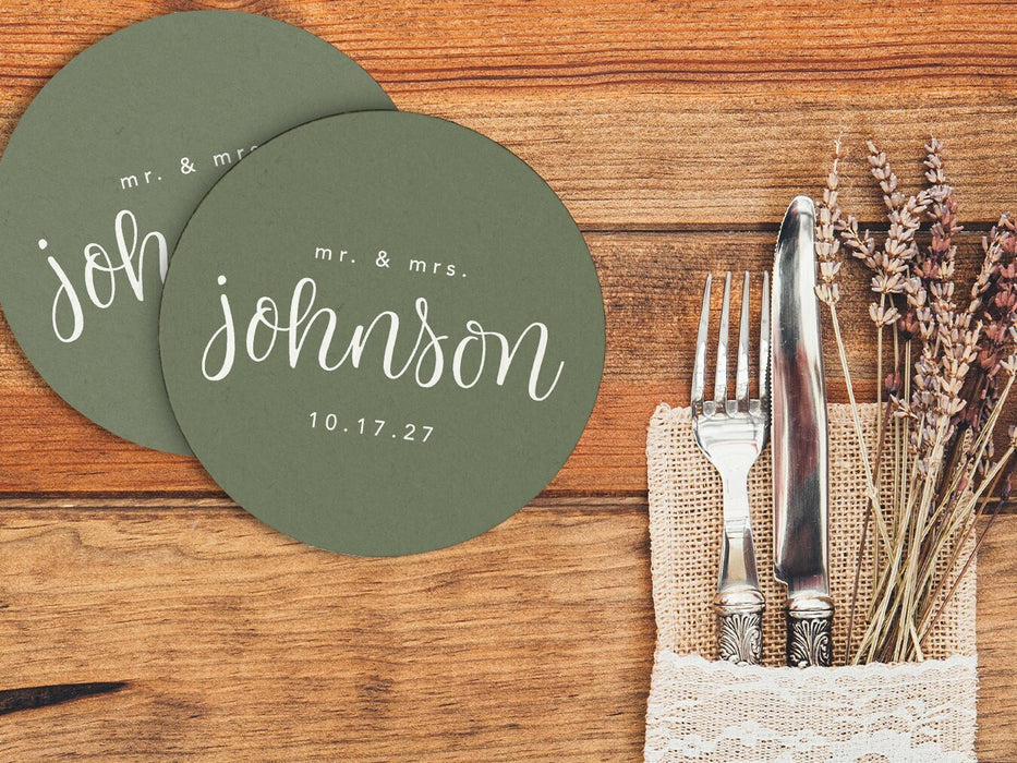 Two coasters are shown next to a place setting and silverware. Coasters feature a custom last name design with a married couple's last name and wedding date.