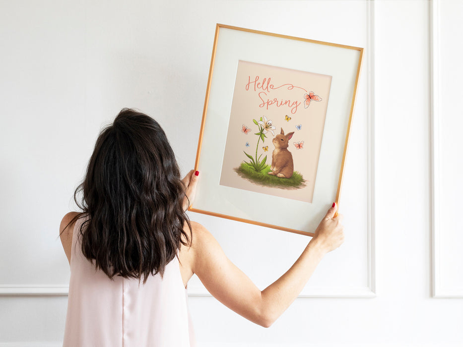 woman with black hair and pink shirt holding up a pastel easter print that says hello spring with a bunny and a lily surrounded by colorful butterflies in a wooden frame