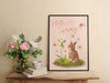 pastel easter print that says hello spring with a bunny and a lily surrounded by colorful butterflies in a black frame on a wooden table next to flowers in a vase as well as a stack of books with a ceramic tea cup sitting on top