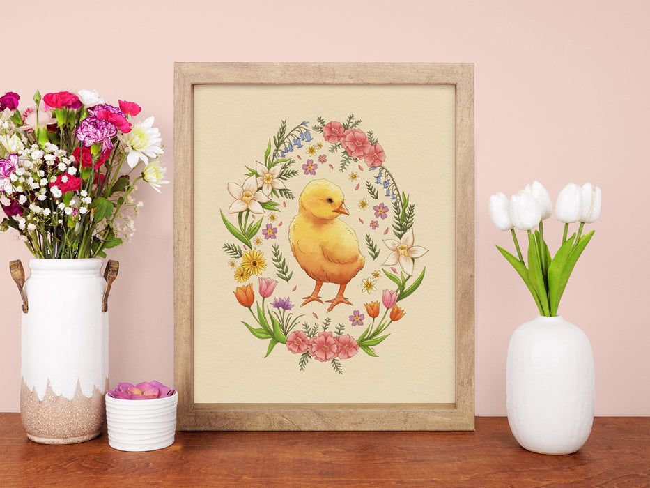 wooden frame with easter print of a baby chick surrounded by spring flowers on a wooden table in front of a pink wall surrounded by colorful potted flowers