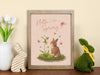 pastel easter print that says hello spring with a bunny and a lily surrounded by colorful butterflies in a wooden frame on a wooden counter neighboring tulips in a vase as well as easter bunny sculptures