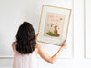 woman with black hair and pink shirt holding up a wooden frame with a pastel easter print that says hello spring with a bunny and a lily surrounded by colorful butterflies