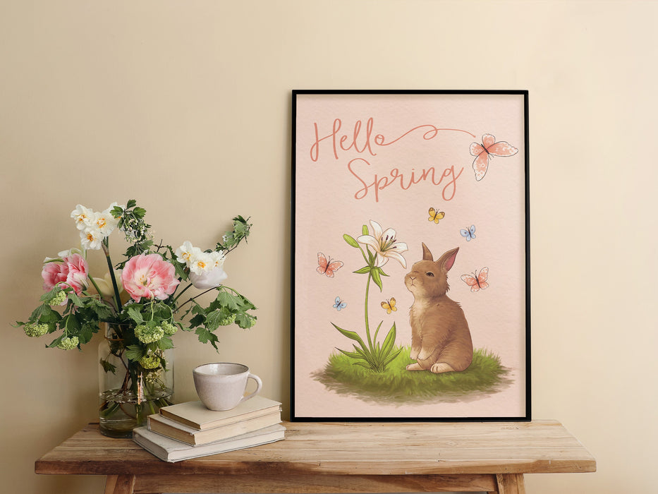 pastel easter print that says hello spring with a bunny and a lily surrounded by colorful butterflies in a black frame on a wooden table next to flowers in a vase as well as a stack of books with a ceramic tea cup sitting on top