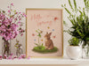 pastel easter print that says hello spring with a bunny and a lily surrounded by colorful butterflies in a gold frame sitting ontop of a white counter surrounded by potted plants and flowers in vases