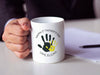 man in a business suit holding a white ceramic mug that says happy first fathers day love Elijah with a yellow handprint design on a white desk next to some papers and a pen