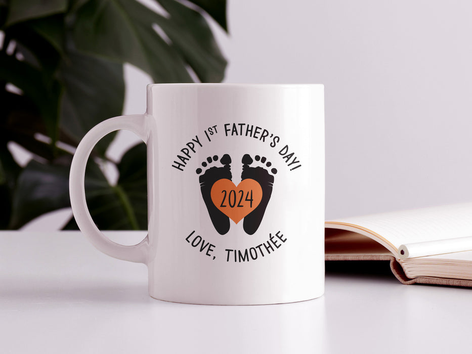 white ceramic mug that says happy first fathers day love Timothee with a footprint design with an orange heart with the year 2024 in it on a white desk next to an open book in front of a house plant