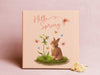 12x12 canvas with Hello Spring Easter print with a bunny and butterflies in front of a purple wall with small flowers in front of the canvas