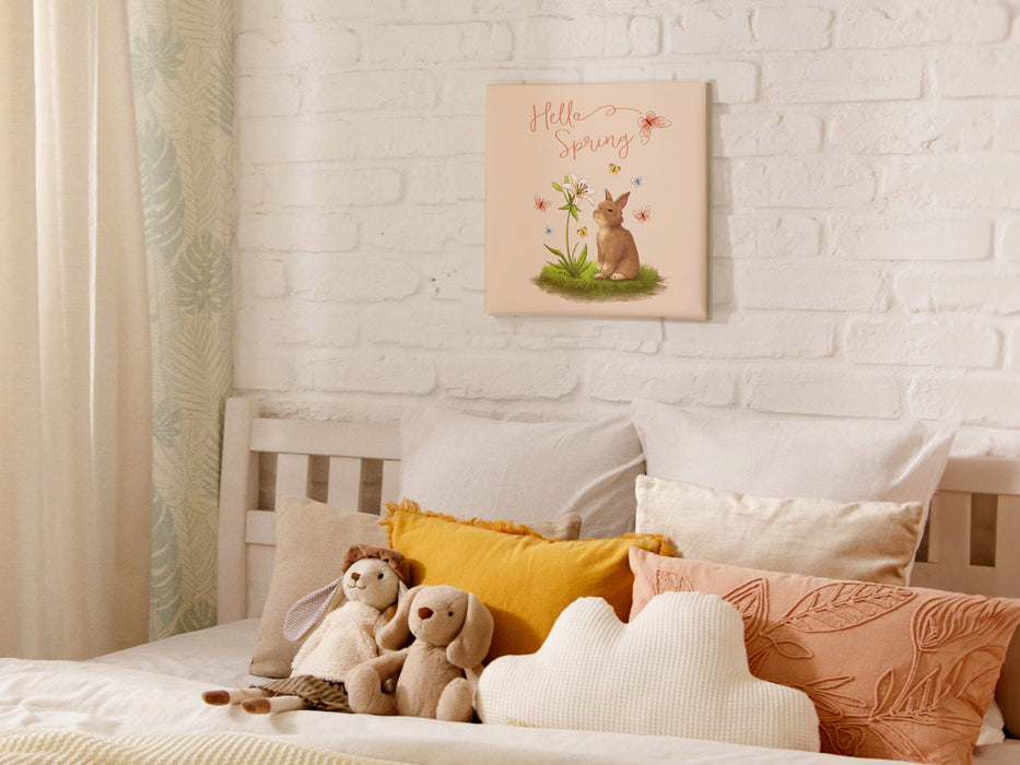12x12 canvas with Hello Spring Easter print with a bunny and butterflies on white brick wall hanging over bed in bedroom decorated with cute pillows and rabbit stuffed animals