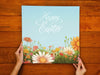 two hands holding a 12x12 canvas of happy easter artwork of a spring meadow on a wooden table