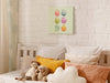 12x12 canvas with colorful easter artwork of rows of eggs and a baby chick hanging over a bed with multicolored pillows and stuffed rabbits in a room that has plant patterned curtains and a white brick wall