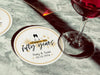 A single coaster with a wine glass on top of it with a coaster off to the side. Coasters shown are customizable. Coasters are designed with gold sparkly elements, wine glasses, and the words Cheers to twenty-five years, custom names, and custom date.