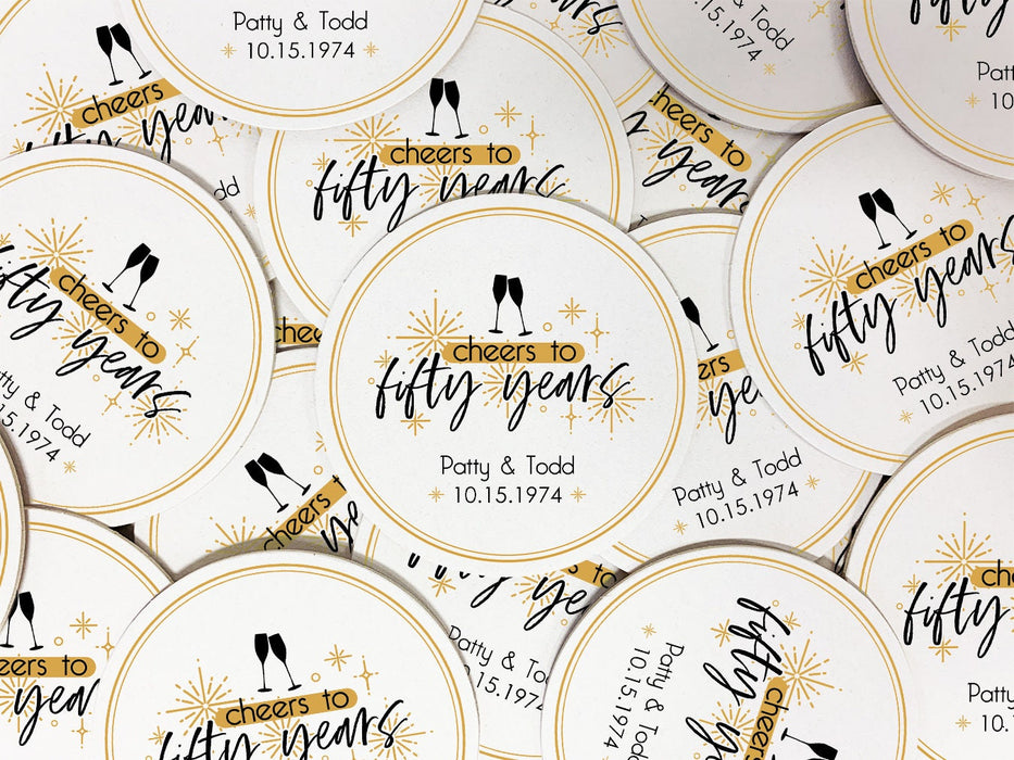 Coasters are shown scattered on a flat surface. Coasters shown are customizable. Coasters are designed with gold sparkly elements, wine glasses, and the words Cheers to twenty-five years, custom names, and custom date.