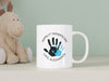 white ceramic mug that says happy first fathers day love Alexander with a blue handprint design on a white counter next to a dinosaur stuffed animal