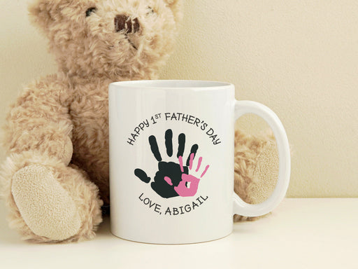 white ceramic mug that says happy first fathers day love Abigail with a pink handprint design on a white counter next to a brown stuffed teddy bear