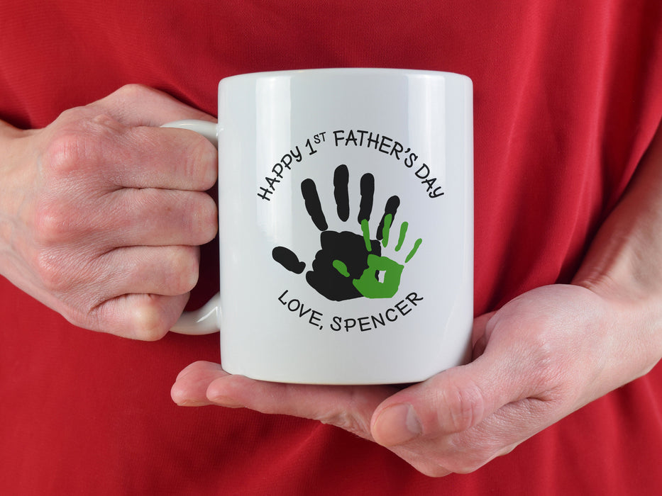 man in a red shirt holding a white ceramic mug that says happy first fathers day love Spencer with a green handprint design