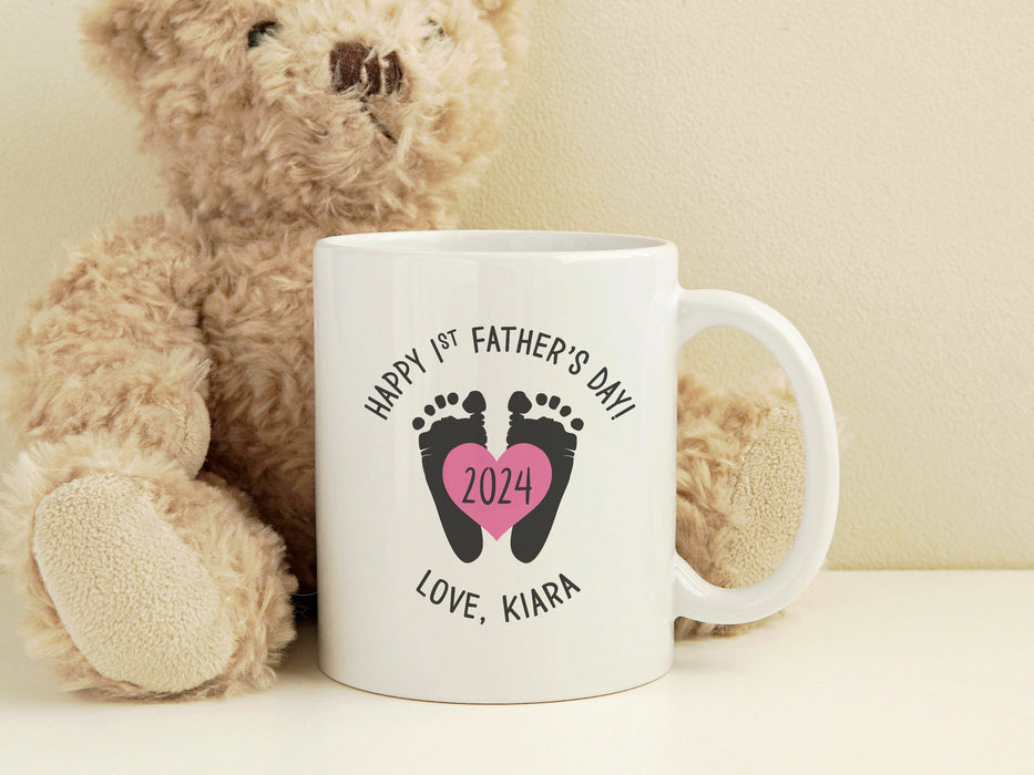 a white ceramic mug that says happy first fathers day love Kiara with a footprint design with a pink heart with the year 2024 in it on a white surface next to a brown stuffed teddy bear