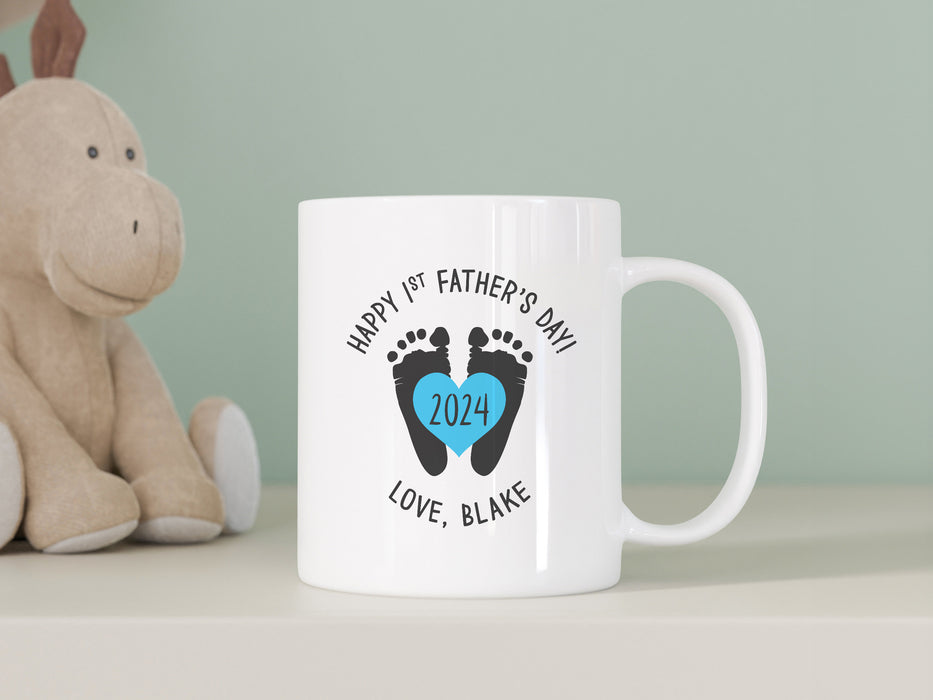 white ceramic mug that says happy first fathers day love Blake with a footprint design with a blue heart with the year 2024 in it on a white counter next to a dinosaur stuffed animal