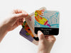 two hands holding four square paper coasters with kids artwork and custom text printed on the coasters against a white background