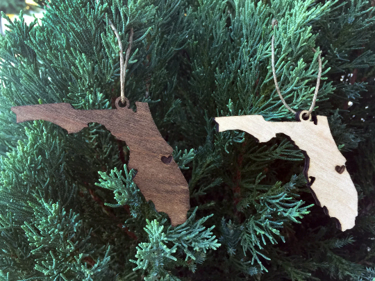 Ornaments shown hanging from Christmas tree. These ornaments are made out of wood and shaped as Florida. A heart is laser etched where chosen hometown or city would be.